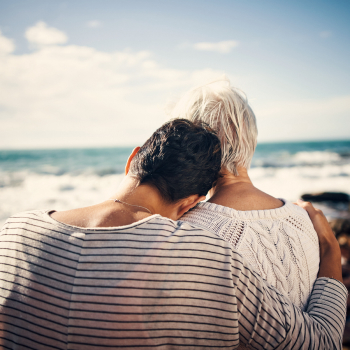 A woman on a beach with her arm around her mother, their backs to the camera as they look out across the sea.